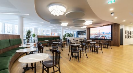 Une brasserie parisienne a été installée dans des locaux professionnels. Des chaises, des bancs verts, des tables en marbres et des plantes sont disposés un peu partout dans la pièce.