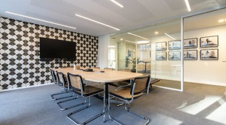 Salle de réunion qui accueille une table de réunion et huit chaises noires Marcel Breuer. Un mur est décoré d'!un papier peint vintage et au milieu est disposé un grand écran télé.
